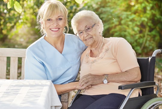 A smiling caregiver sits beside an elderly woman in a wheelchair outdoors, both holding hands and looking at the camera. Sunlight filters through green trees in the background, creating a warm, cheerful atmosphere.