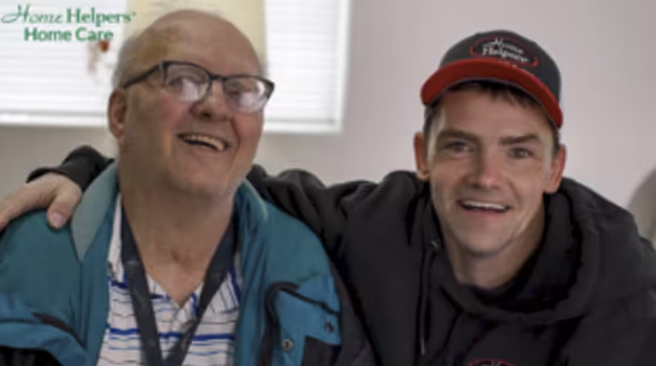 Two smiling men, one older wearing glasses and a striped shirt under a blue jacket, the other younger in a black hoodie and cap, sit close together indoors. Home Helpers Home Care logo is visible in the top left corner.