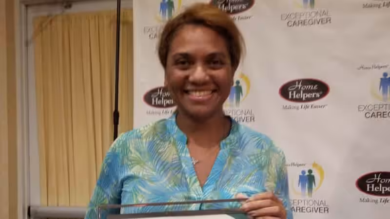 A woman smiles while holding a certificate in front of a backdrop that reads Home Helpers and Exceptional Caregiver. She is wearing a blue and green patterned blouse.