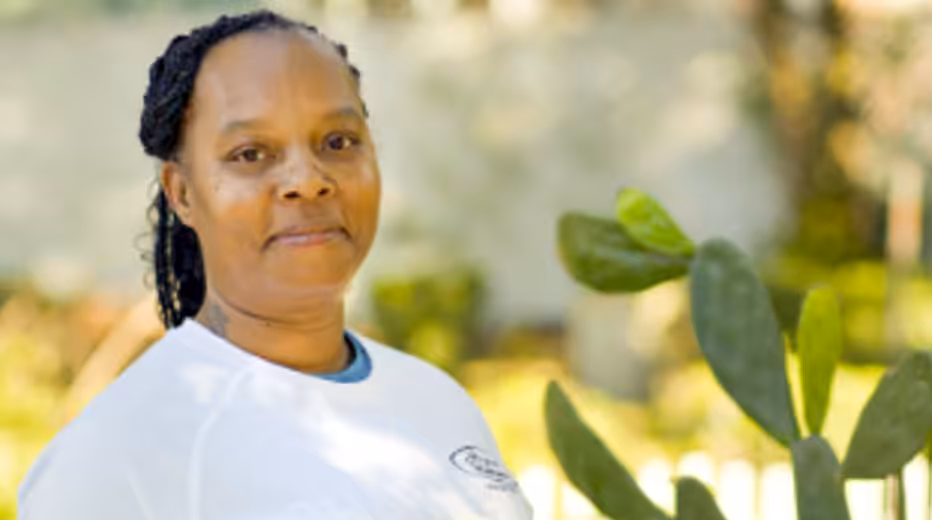 A woman with braided hair stands outdoors, wearing a white shirt. She is looking at the camera and smiling slightly. Green plants and a blurred fence are visible in the sunlit background.
