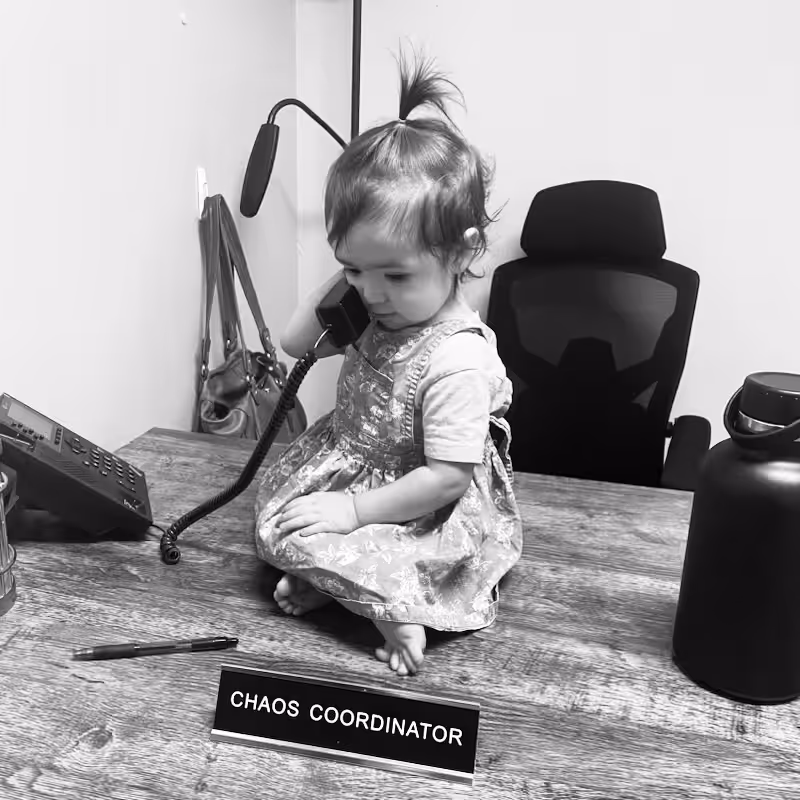 A toddler in a dress sits on an office desk, holding a phone to her ear. A nameplate reading CHAOS COORDINATOR is in front of her, along with a pen, a water bottle, and an office chair in the background.
