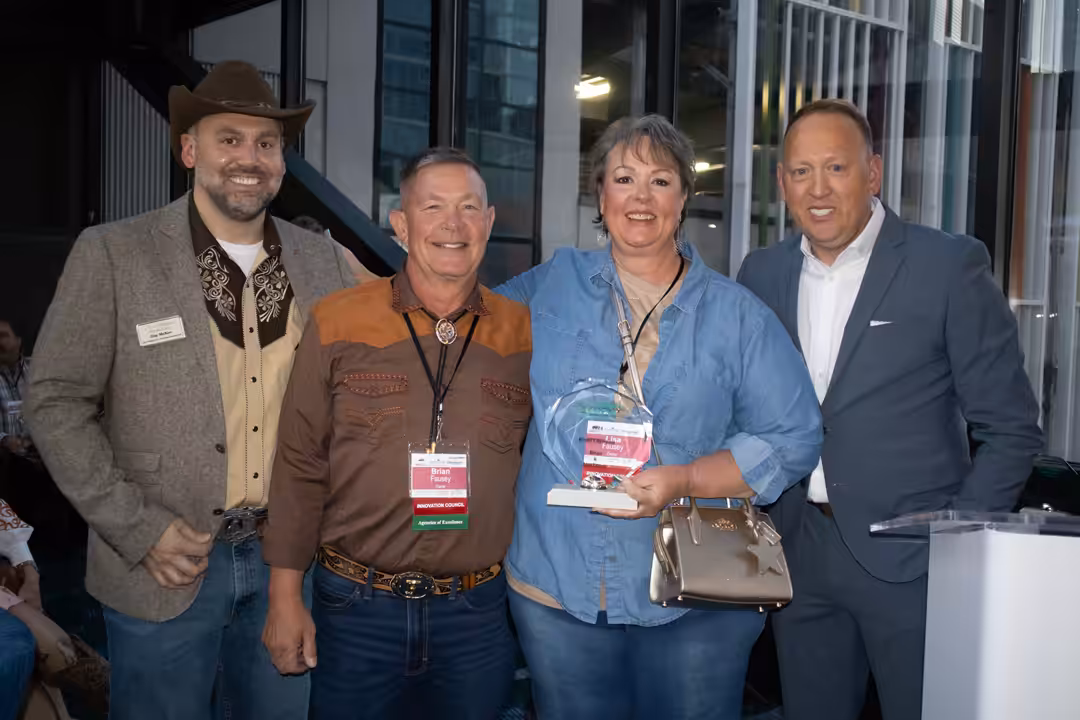 Four adults pose together indoors at an event. One woman holds a glass award and a small purse. The three men stand beside her, smiling. Two men wear casual western attire; one wears a suit. Name badges are visible on their chests.