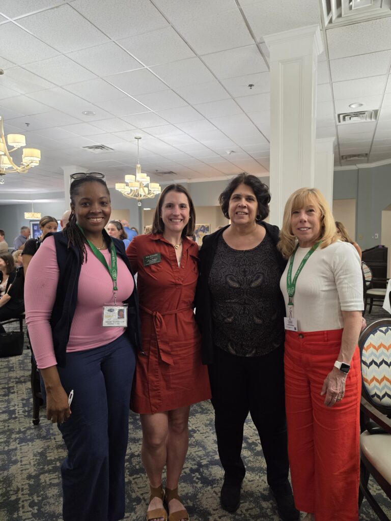 Four women stand together, smiling for a group photo at an indoor event. They wear name badges and casual business attire. Other people and tables are visible in the bright, conference-style room behind them.