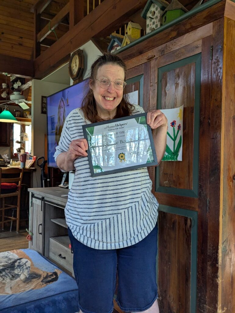 A smiling woman in glasses holds a certificate in a cozy, wooden room with art on the wall and a dog lying on a blanket. She wears a striped shirt and denim shorts.