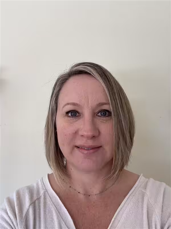 A woman with shoulder-length blonde hair and blue eyes, wearing a light-colored top and a delicate necklace, stands in front of a plain off-white background, looking directly at the camera.