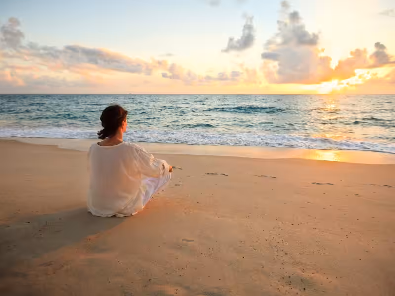 A person in a white dress sits on the sandy beach, facing the ocean and watching the sunrise or sunset, with gentle waves and clouds in the sky.