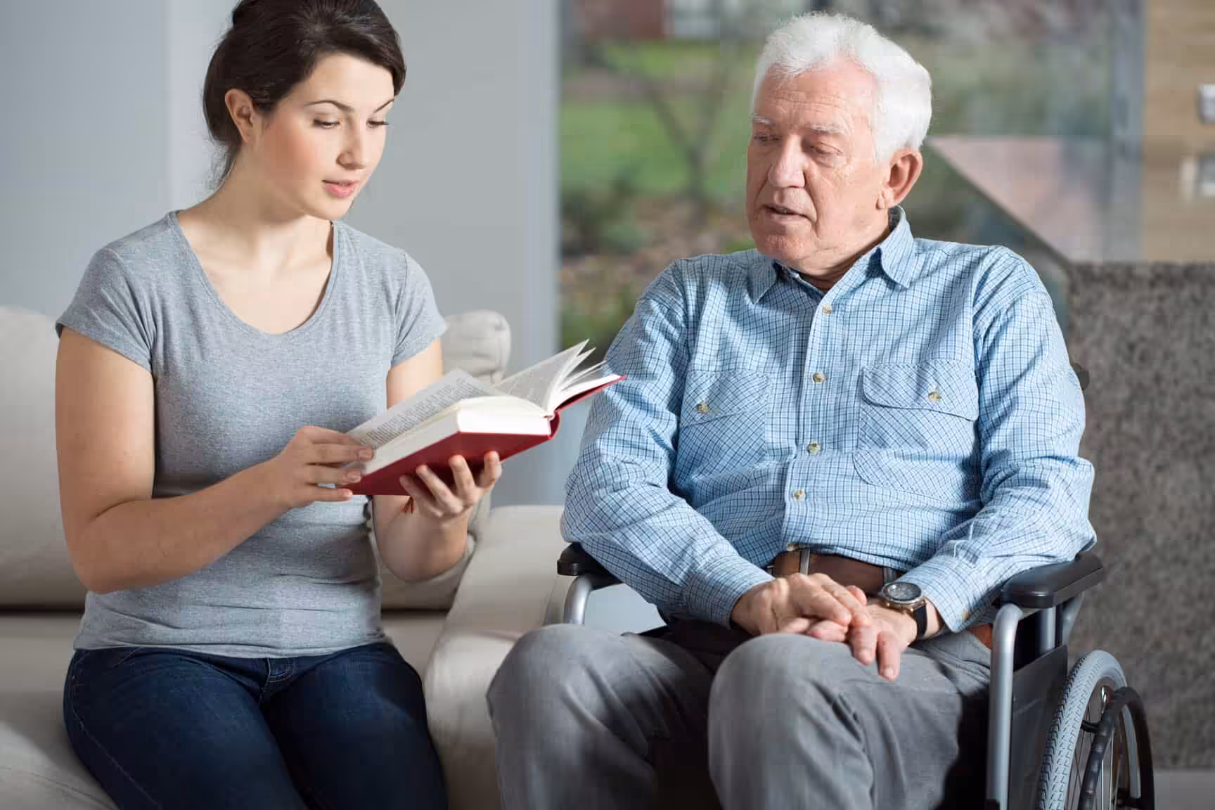 A young woman sits on a couch reading a book to an older man in a wheelchair. Both are indoors, and the man listens attentively while the woman holds the open book and reads.