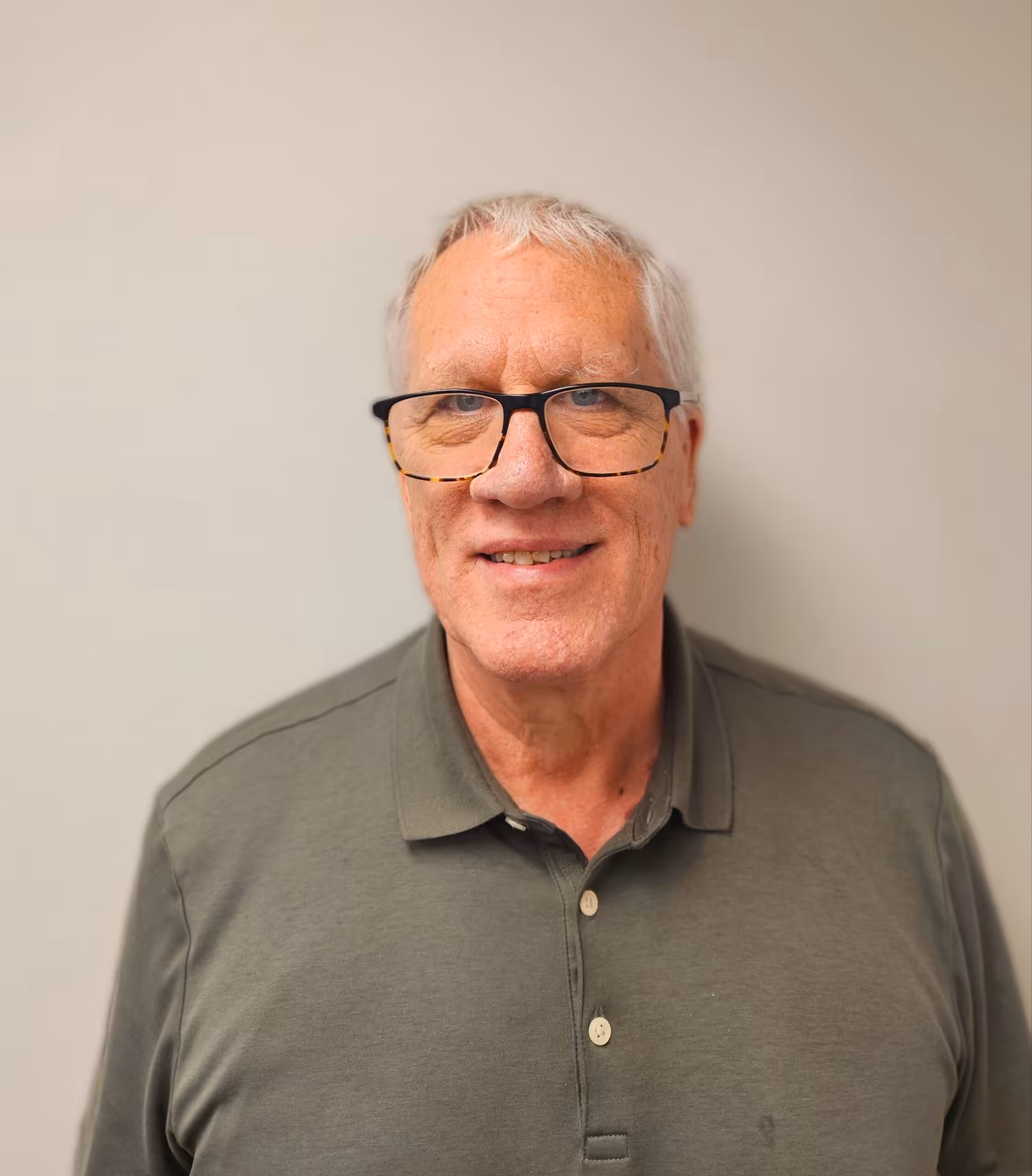 An older man with short gray hair and glasses, wearing a green polo shirt, smiles at the camera against a plain light-colored background.