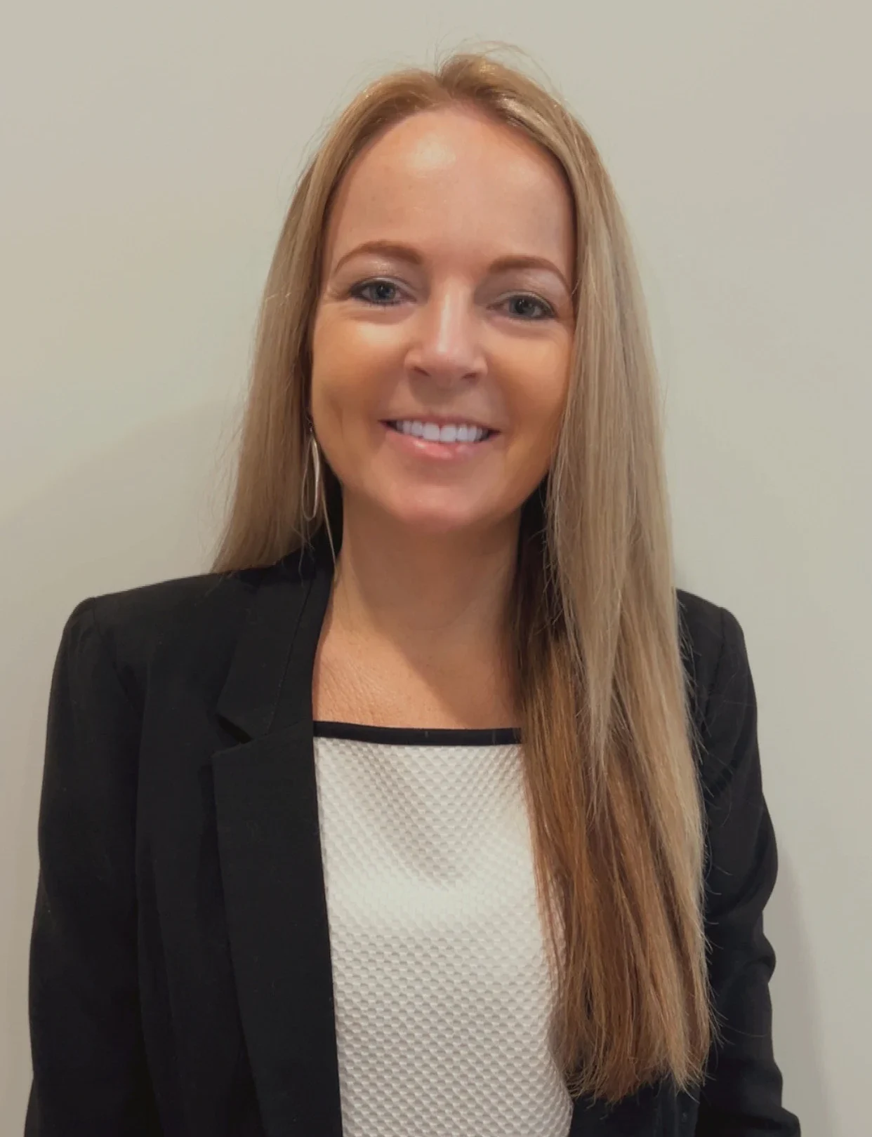 A woman with long straight blonde hair, wearing a black blazer over a white textured blouse, smiles at the camera in front of a plain light-colored background.