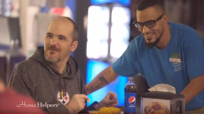 A man in a gray hoodie sits at a table with food and a Pepsi bottle. Another man wearing glasses and a blue shirt stands beside him, smiling. The setting appears to be a casual dining area. The Home Helpers logo is visible.