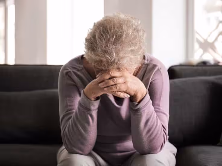 An older adult with gray hair sits on a dark sofa, leaning forward with their head in their hands, appearing distressed or upset. Light filters through windows in the background.
