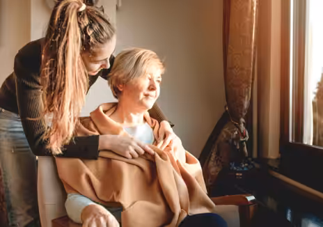 A young woman stands behind an older woman seated in a chair, gently embracing her shoulders as they both look out a sunlit window, sharing a warm and comforting moment together.