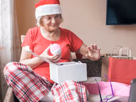 An older woman in a Santa hat and plaid pajamas sits on a couch, smiling as she holds a pink ornament from an open gift box, with shopping bags and eyeglasses nearby.