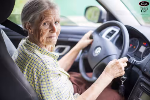 An elderly woman with gray hair, wearing a yellow plaid shirt, sits in the driver’s seat of a car and looks at the camera while holding the steering wheel and car keys.