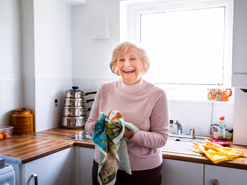 An elderly woman with short blonde hair smiles while drying dishes with a tea towel in a bright, modern kitchen. Sunlight streams through the window behind her, illuminating the countertop and sink area.