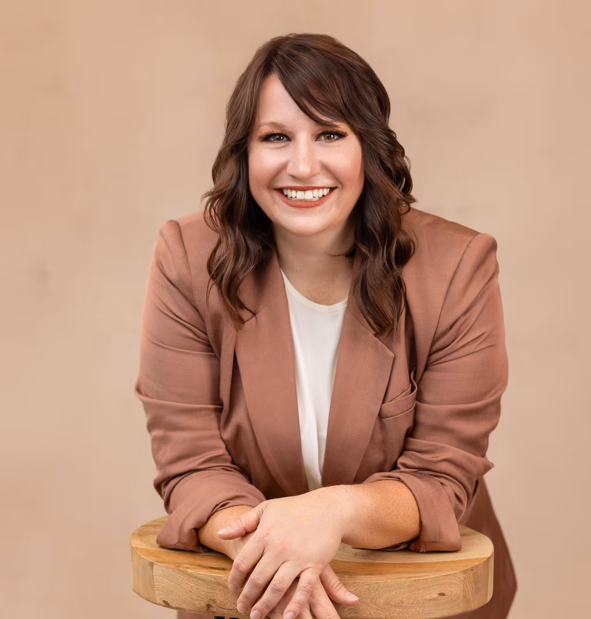 A woman with wavy brown hair, wearing a mauve blazer over a white top, smiles while leaning on a wooden stool against a beige background.