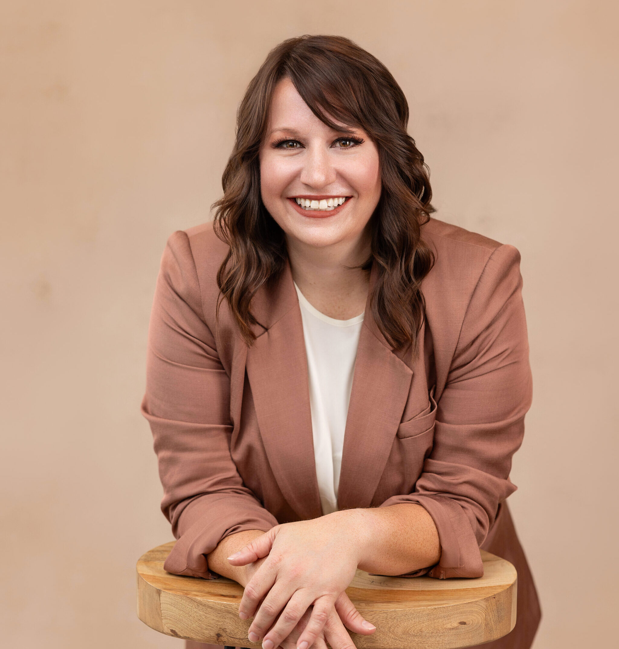 A woman with wavy brown hair, wearing a mauve blazer over a white top, smiles while leaning on a wooden stool against a beige background.
