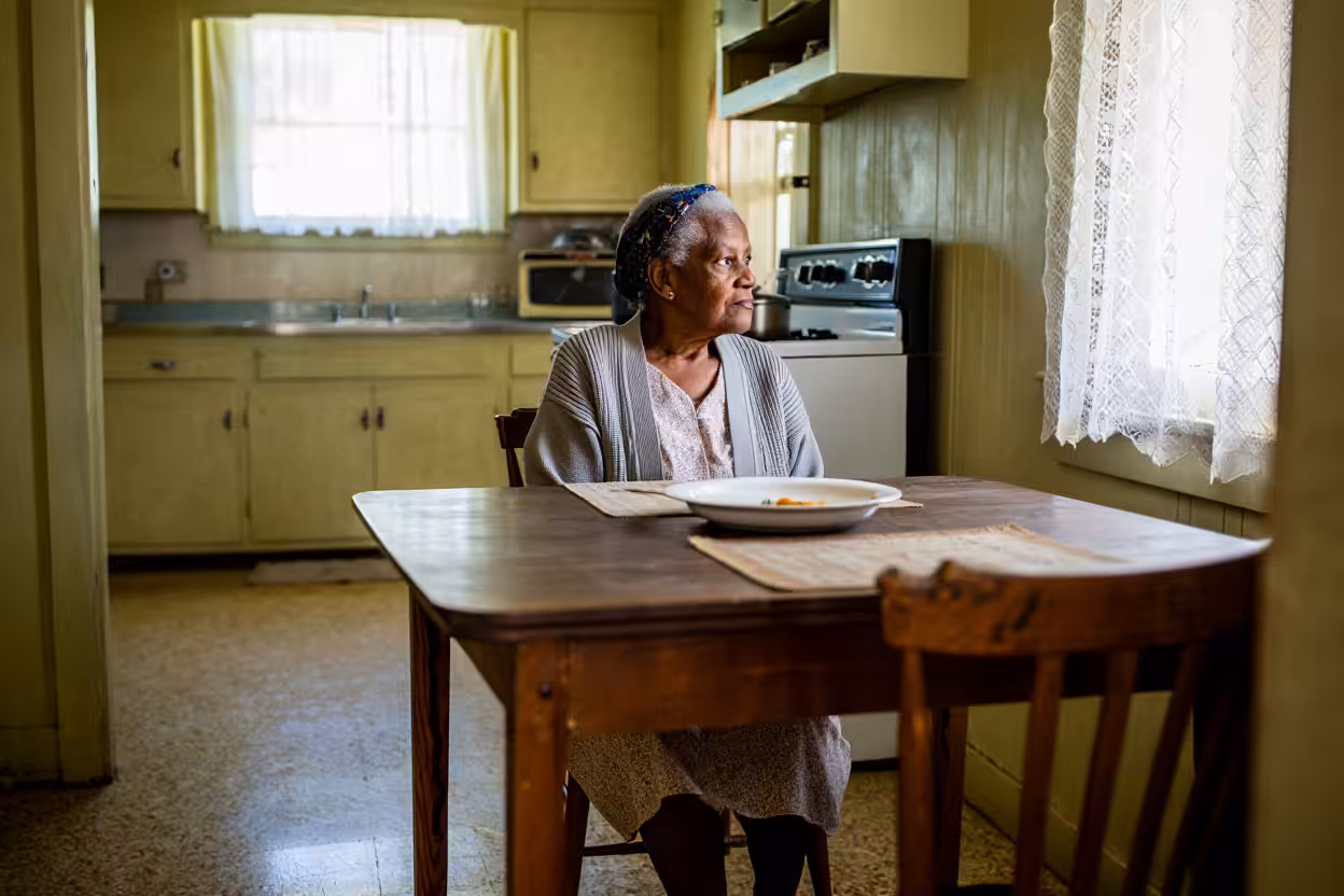 An elderly woman sits alone at a wooden kitchen table, gazing out a sunlit window. A plate with some food is in front of her. The kitchen is modest, with yellow cabinets and lace curtains.