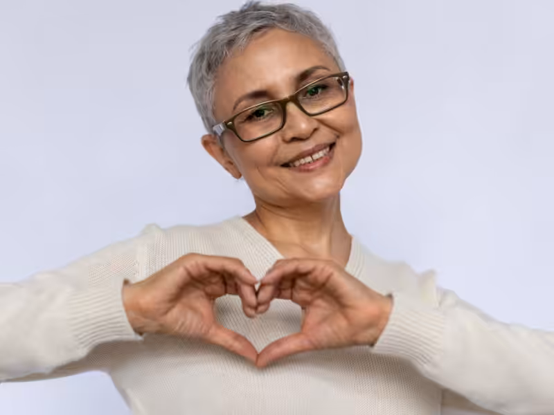 Smiling older woman with short gray hair and glasses making a heart shape with her hands, wearing a light-colored sweater against a plain light background.