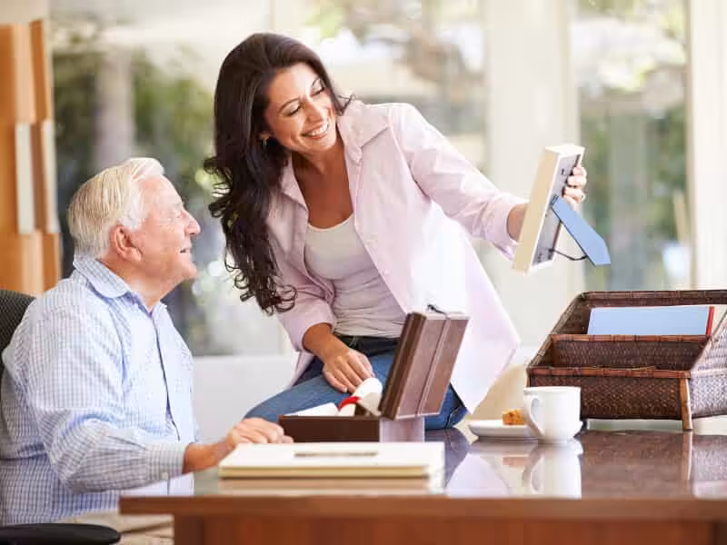 A smiling woman shows a framed photo to an elderly man while sitting at a table with an open box, papers, and a coffee cup, suggesting they are reminiscing or organizing memories together.