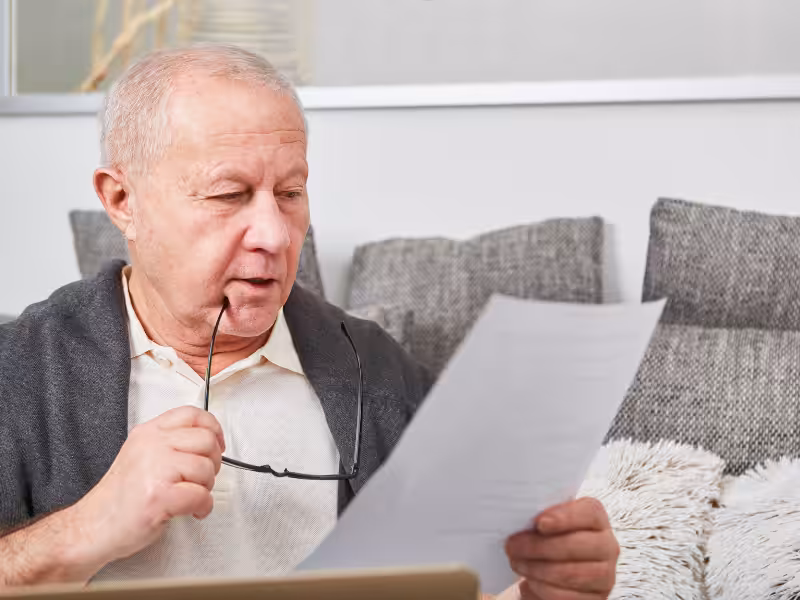 An older man sitting on a sofa holds his glasses and looks intently at a sheet of paper, appearing focused and thoughtful. Cushions and a laptop are visible in the background.