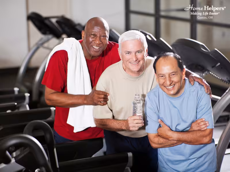Three smiling older men pose together at a gym, standing in front of treadmills. One man holds a water bottle, another has a towel draped over his shoulder. They appear happy and relaxed after exercising.