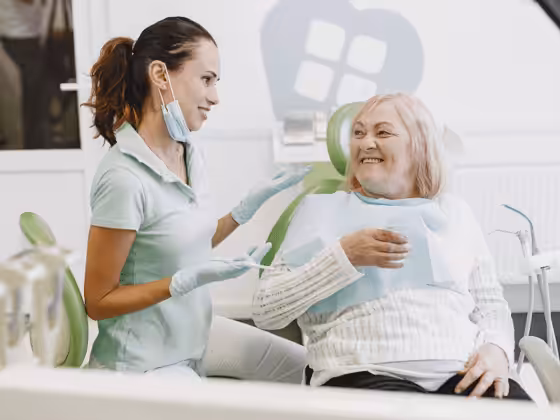 A dental professional in scrubs and gloves talks with an older woman sitting in a dental chair, both smiling. The patient wears a dental bib, and the setting appears to be a modern dental clinic.