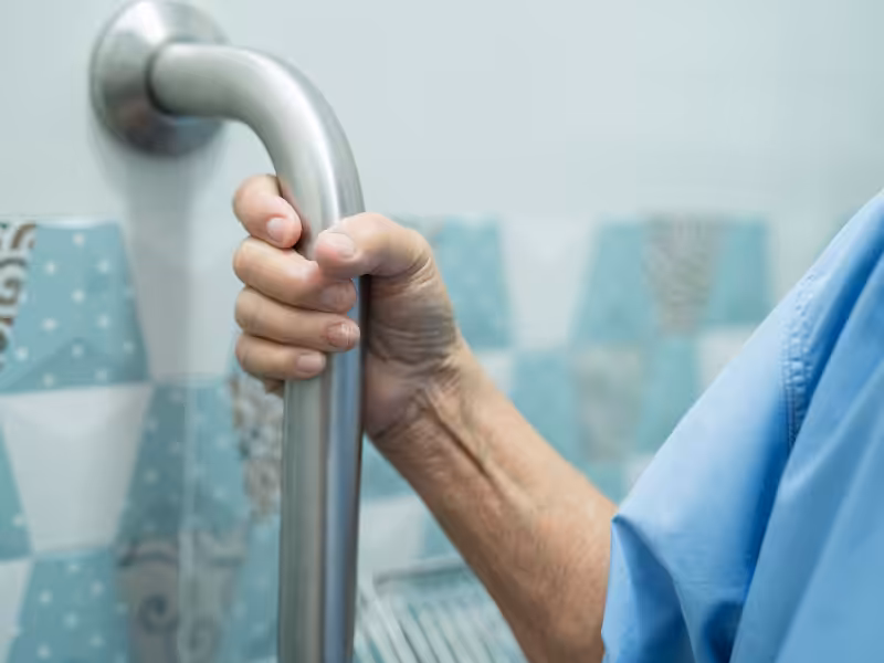 A person wearing a blue shirt grips a metal handrail in what appears to be a bathroom, with blue and white tiles visible in the background.