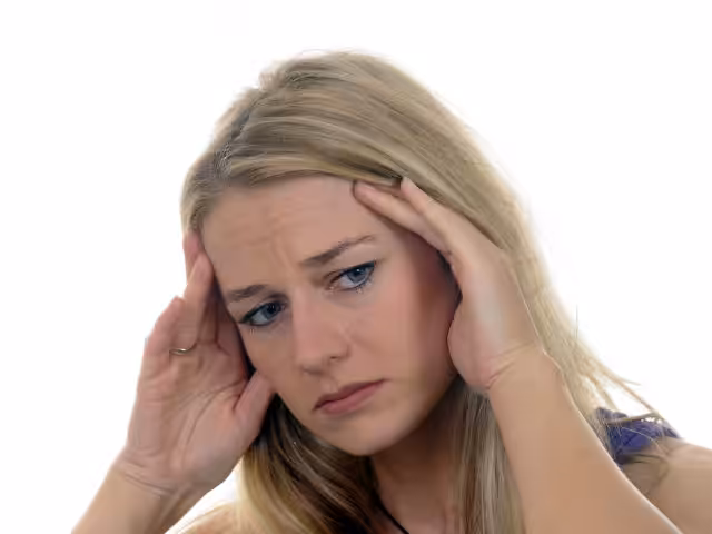 A woman with long blonde hair holds her head with both hands, appearing stressed or worried, against a plain white background.