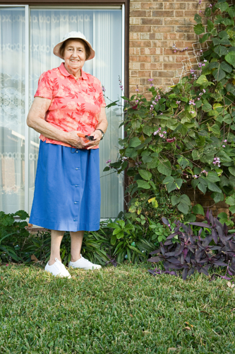 An older woman in a hat, red floral shirt, and blue skirt stands on a lawn beside a brick house with a window and flowering plants. She is smiling and holding something in her hands.