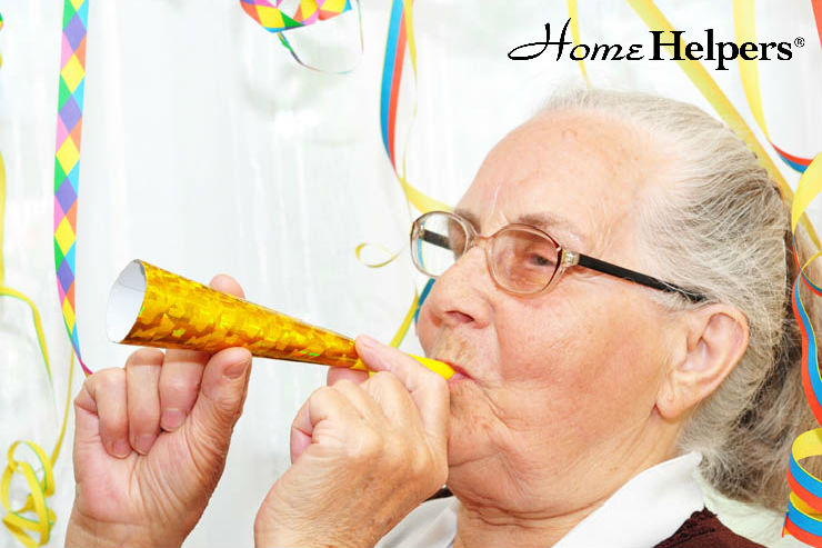 An elderly woman with glasses blows a yellow party horn, surrounded by colorful streamers, with the Home Helpers logo in the top right corner.