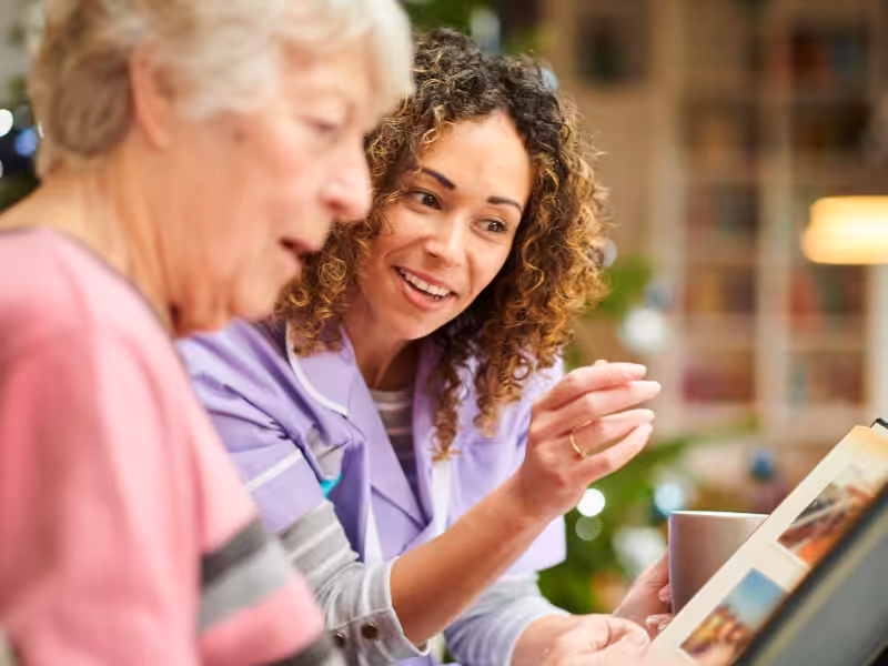 A young caregiver in a purple uniform smiles while helping an older woman look through a photo album indoors, suggesting a warm, supportive interaction.