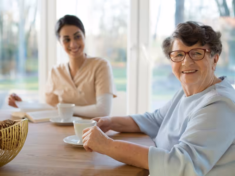 An older woman with glasses smiles while holding a cup at a table, with a younger woman sitting behind her, also smiling. They appear to be enjoying a conversation over coffee in a bright room.