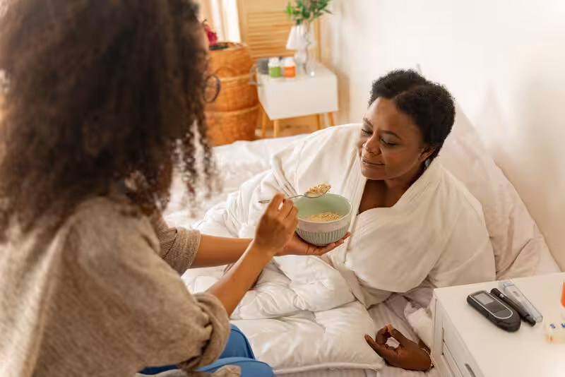 A woman in bed, wearing a robe, is being fed oatmeal from a bowl by another person. Medical supplies, including a glucose meter, are on the bedside table.
