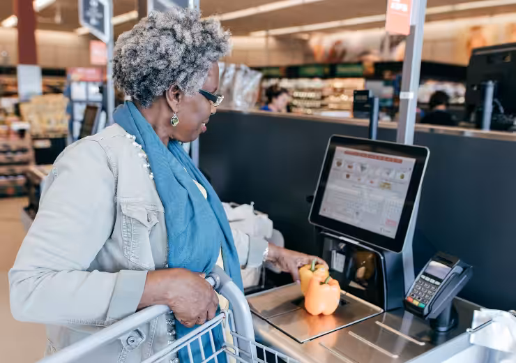 An older woman scans a yellow bell pepper at a self-checkout kiosk in a grocery store, holding a shopping cart and wearing glasses, earrings, and a blue scarf.