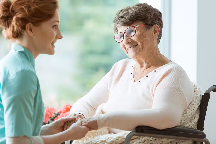 A smiling elderly woman in a wheelchair holds hands with a caregiver who is kneeling beside her, both sharing a warm moment near a window.