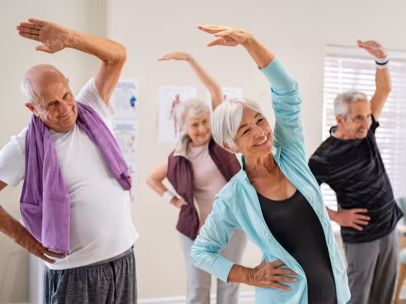 Four older adults in casual workout clothes smile while performing a group exercise, standing and stretching with one arm overhead in a bright room with fitness posters on the wall.