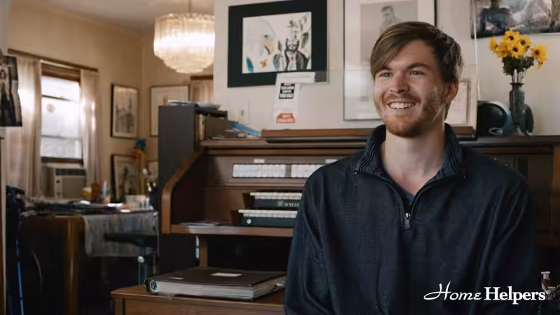 A smiling man with short brown hair and a beard sits indoors near a cluttered desk and flowers. Art and photos decorate the wall behind him. The Home Helpers logo appears in the bottom right corner.