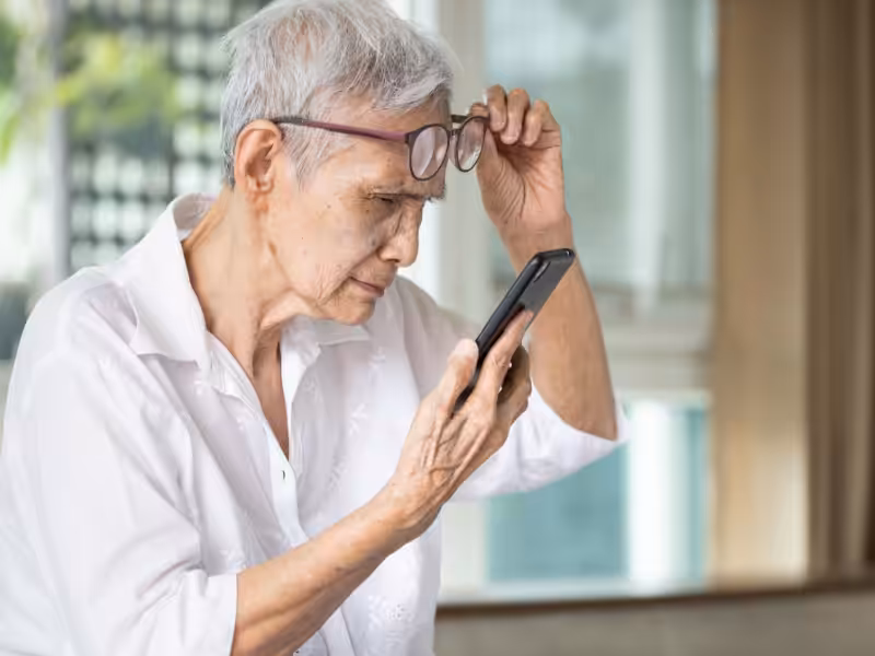 An older adult with short gray hair holds their glasses up and looks closely at a smartphone, appearing to focus carefully on the screen in a brightly lit room.