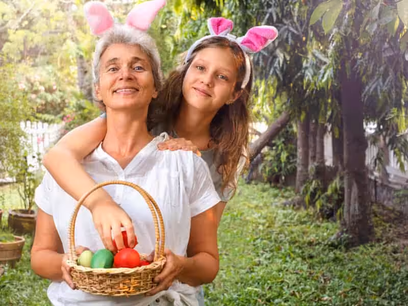 Two people wearing bunny ears smile together outdoors. One holds a basket filled with colorful eggs while the other hugs from behind. They are surrounded by greenery in a garden setting.