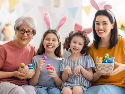 Four smiling people, including an older woman, two young girls, and a younger woman, wear bunny ears and hold colorful Easter eggs in baskets, celebrating together indoors.