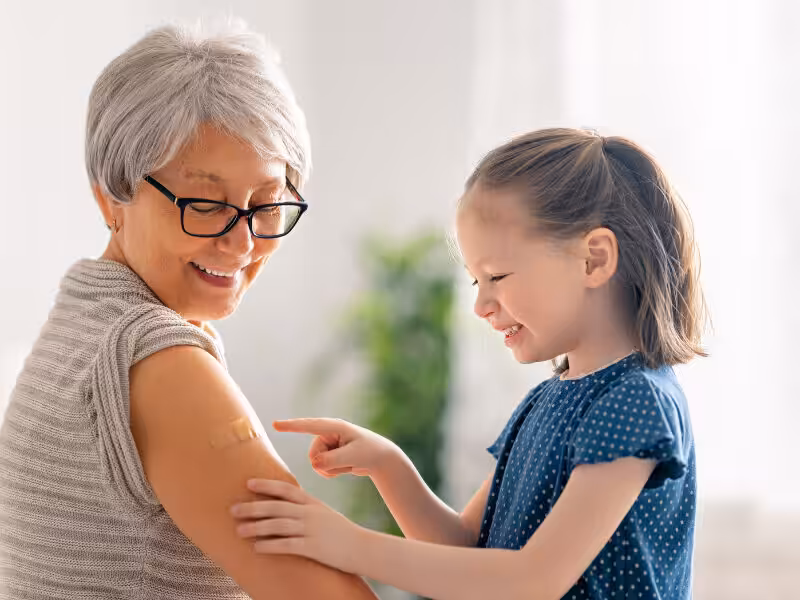 A smiling older woman with gray hair and glasses shows a bandage on her upper arm while a young girl in a blue dress points at it and laughs. They are inside, with soft light in the background.