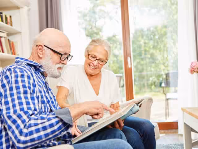 An elderly man and woman sit together on a couch, smiling and looking at a photo album. The man, wearing glasses and a plaid shirt, points at a photo while the woman in a white top looks on happily.