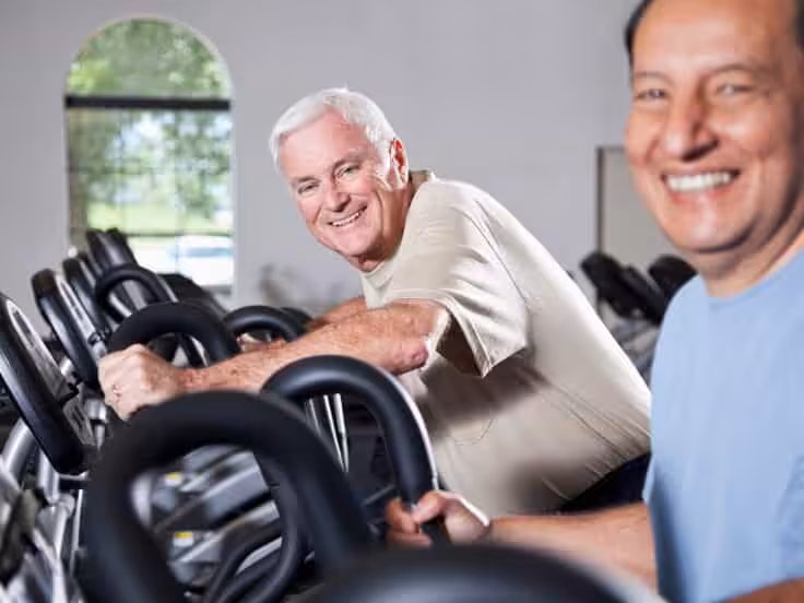 Two older men smiling while using exercise bikes at a gym. One man wears a beige shirt, the other wears blue. Bright room with large windows and multiple exercise machines visible in the background.