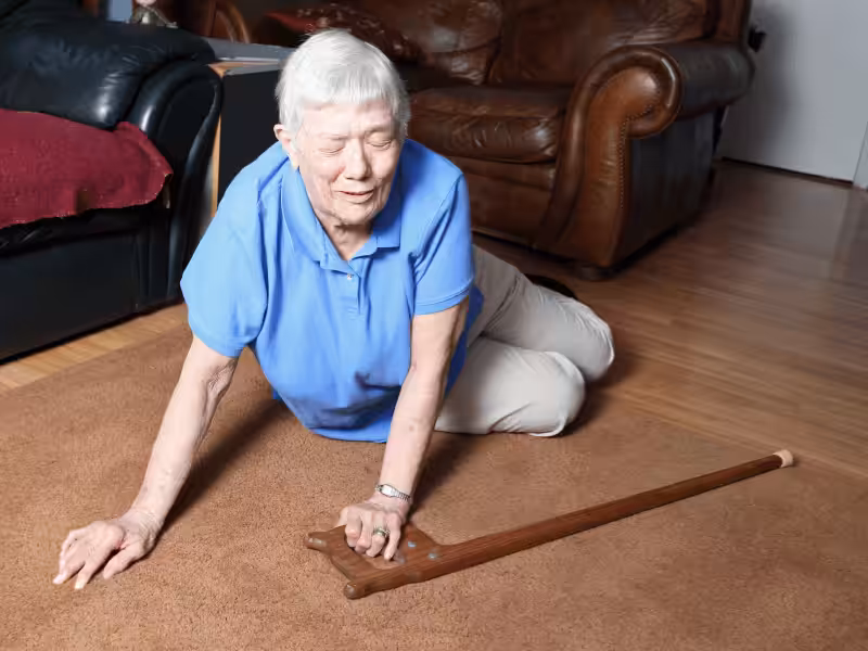 An elderly woman in a blue shirt is on the floor, appearing to have fallen, with her cane lying nearby on a brown carpet in a living room.