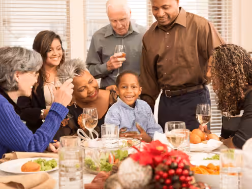 A group of people of various ages gather around a dining table, smiling and enjoying each others company. The table is set with food, drinks, and festive decorations.