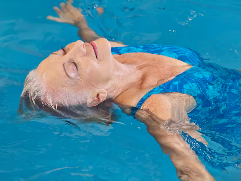 An older woman in a blue patterned swimsuit floats on her back in a swimming pool, eyes closed and smiling peacefully with arms outstretched, enjoying the water.