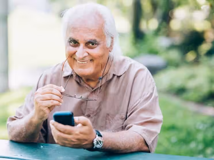 An older man with white hair, wearing a light brown shirt, smiles while holding eyeglasses in one hand and a smartphone in the other, sitting at an outdoor table with greenery in the background.