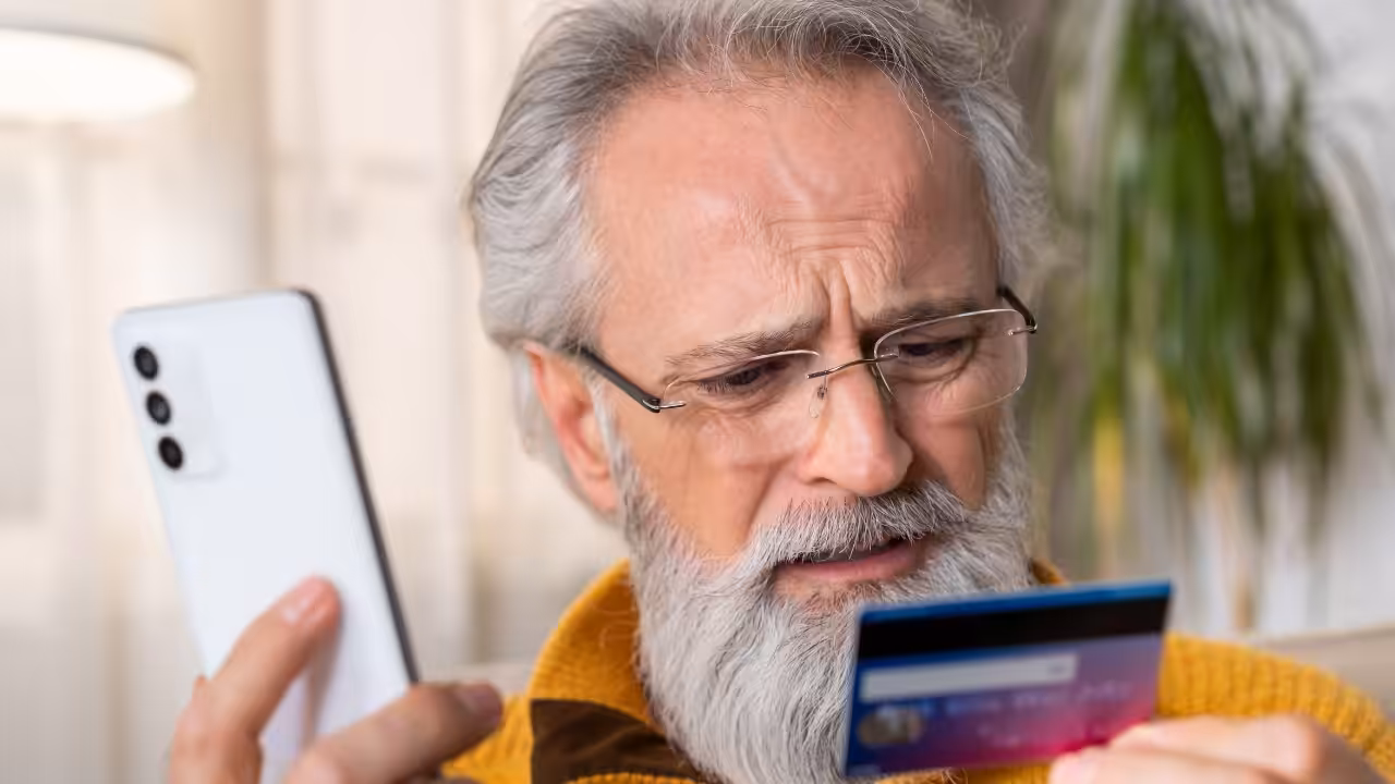 An older man with gray hair and glasses looks worriedly at a credit card in one hand while holding a smartphone in the other, sitting indoors with a blurred background.