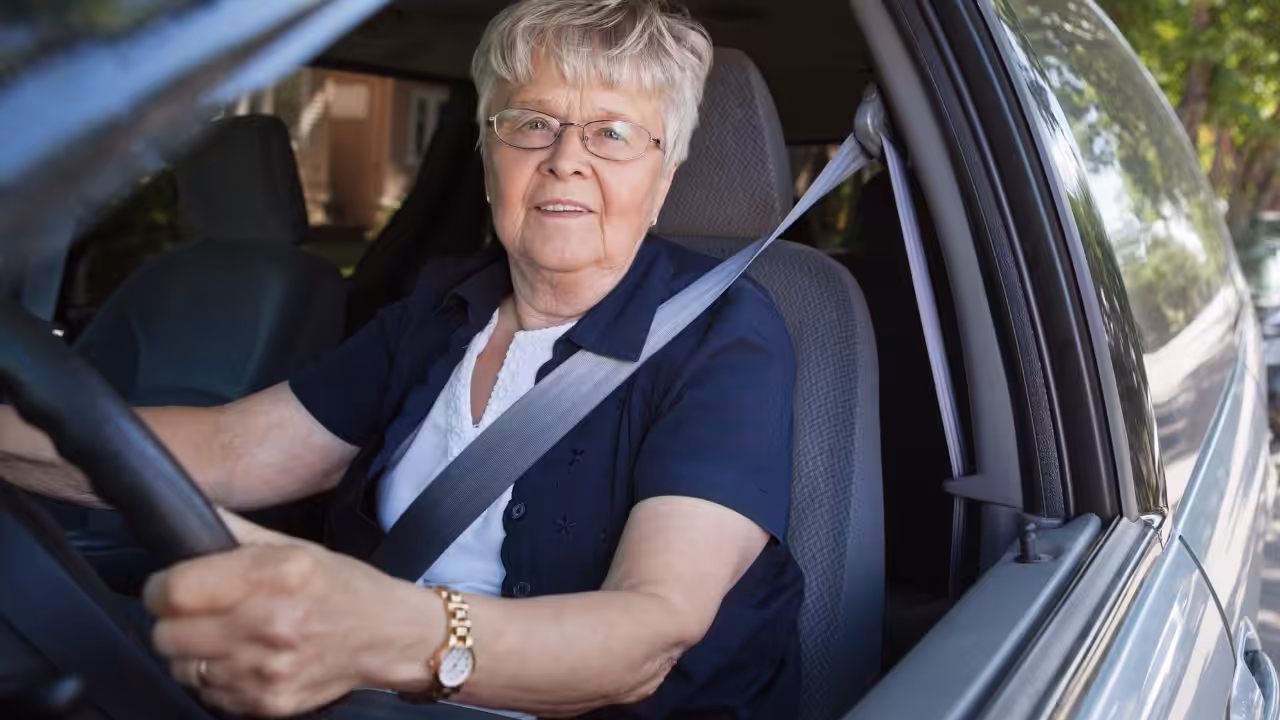 An older woman with short gray hair, glasses, and a white shirt with a dark cardigan is sitting in the driver’s seat of a car, wearing a seatbelt and holding the steering wheel, looking out the window.